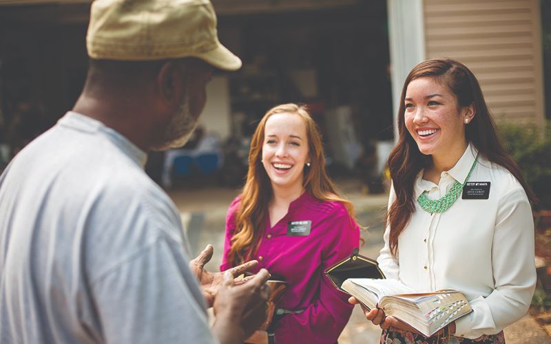 Two Sister Missionaries holding open scriptures and talking to a man outside of his home.