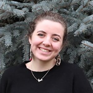 Portrait of a smiling BYU student wearing a black blouse with an evergreen tree as background.